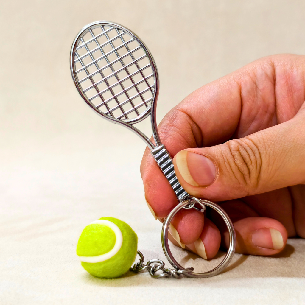 A miniature metal tennis racket keychain with a small yellow tennis ball charm, held by a hand against a light beige background.