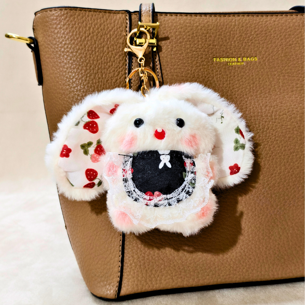 A white faux-fur bunny keychain or bag charm with embroidered strawberry ears, pink cheeks, and a black mouth area, hanging from a gold clasp on a brown textured handbag.