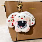 A white faux-fur bunny keychain or bag charm with embroidered strawberry ears, pink cheeks, and a black mouth area, hanging from a gold clasp on a brown textured handbag.