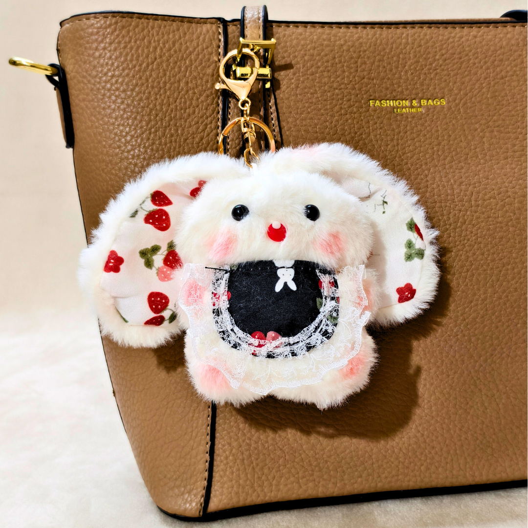 A white faux-fur bunny keychain or bag charm with embroidered strawberry ears, pink cheeks, and a black mouth area, hanging from a gold clasp on a brown textured handbag.