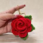 Hand holding a red crocheted rose keychain with a green leaf on a beige background