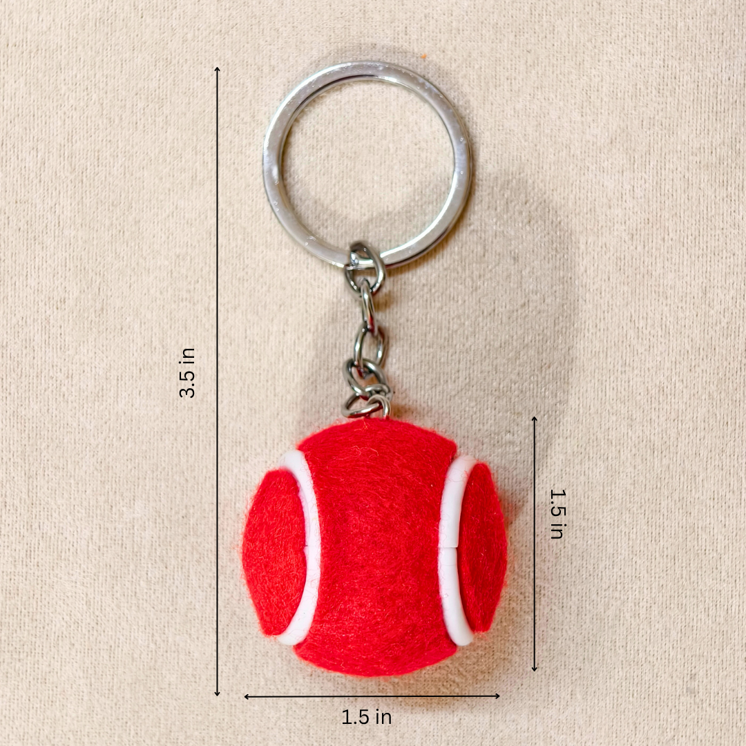 A red felt tennis ball keychain with a silver metal key ring and chain, shown on a beige fabric background with black dimension lines and text indicating measurements of 3.5 in height, 1.