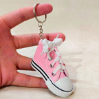 A miniature pink canvas high-top sneaker keychain with white laces and a white rubber toe cap, held in a person's hand against a neutral beige background.