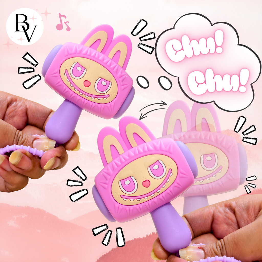 Two hands holding pink and purple bunny-shaped toy rattle mallets with cartoon eyes and teeth, shown against a soft pink background with decorative speech bubbles and motion lines.