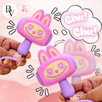 Two hands holding pink and purple bunny-shaped toy rattle mallets with cartoon eyes and teeth, shown against a soft pink background with decorative speech bubbles and motion lines.
