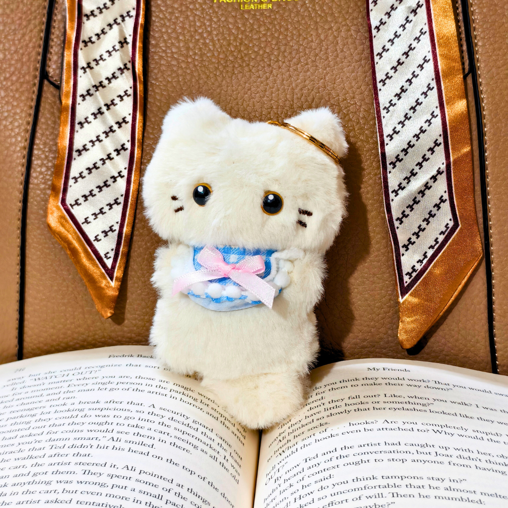 A white plush cat toy with black eyes and a pink bow, positioned between open books with a patterned book spine in the background.