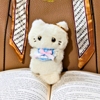 A white plush cat toy with black eyes and a pink bow, positioned between open books with a patterned book spine in the background.