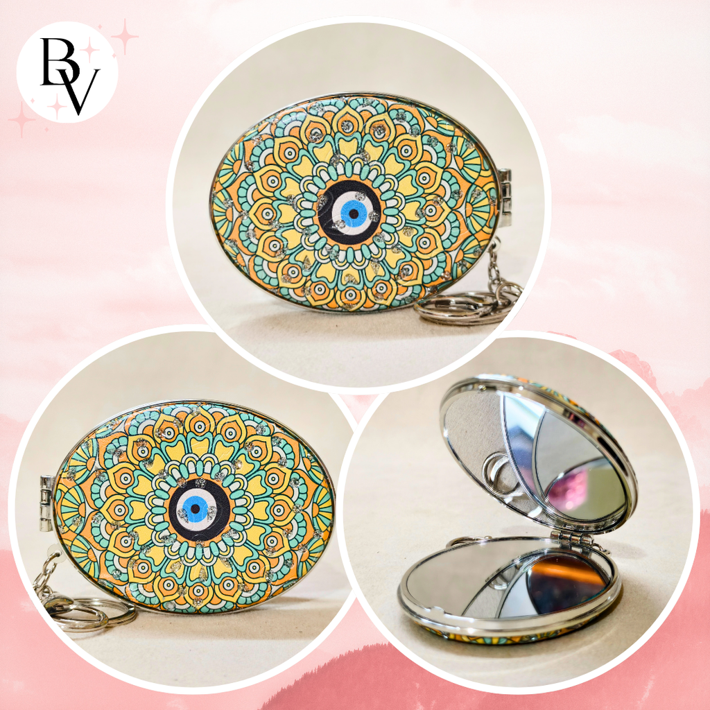 Oval compact mirror keychain with a colorful mandala pattern and blue evil eye design, shown closed and open on a soft pink background.
