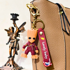 Groot keychain with a 'Lovely baby' tag attached to a brown bag, with a bronze statue in the background.