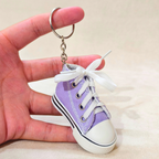 A miniature lavender purple canvas high-top sneaker keychain with white laces and a white rubber toe cap, held in a person's hand against a light beige background.