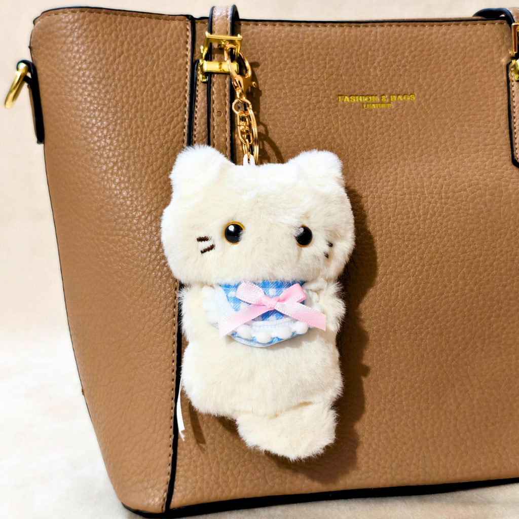 A white plush cat keychain charm with embroidered facial features, a blue-and-white checkered dress, and a pink bow, hanging from a gold ring on a brown textured handbag.