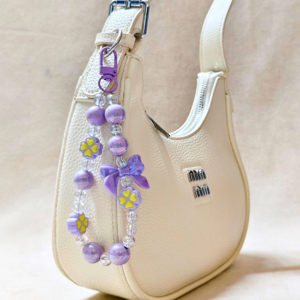 Lilac Bow Charm Attached On Hand Bag In The Front Of Beige Background.