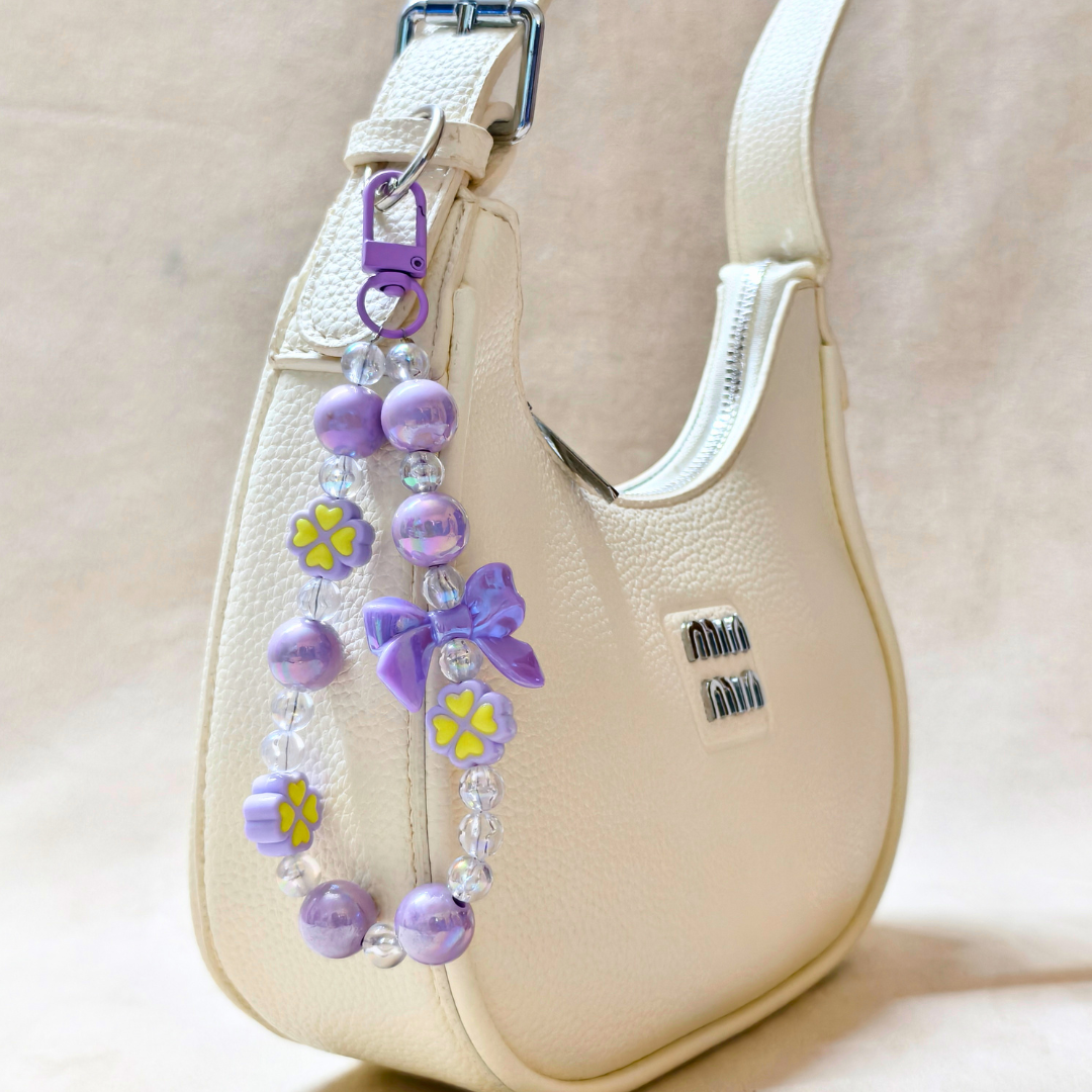 Lilac Bow Charm Attached On Hand Bag In The Front Of Beige Background.