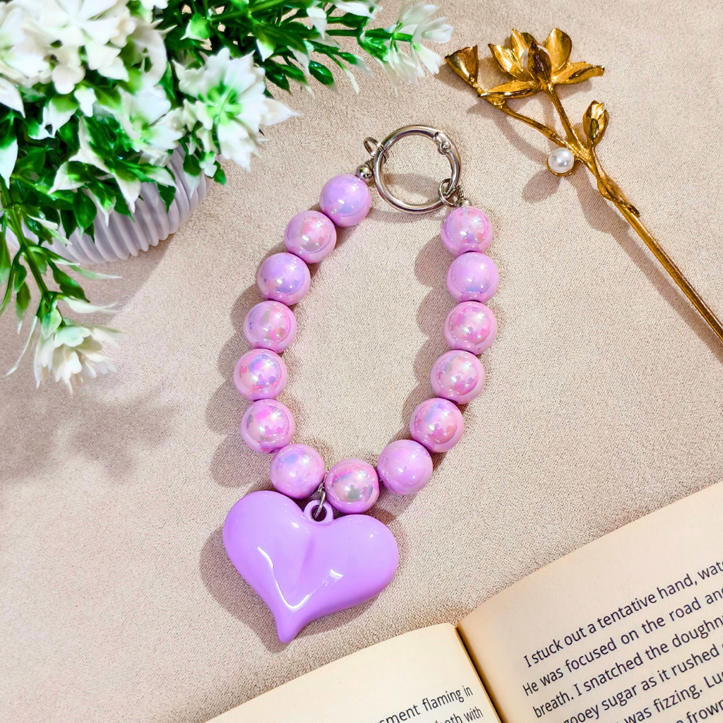 Heart-shaped purple keychain with pink beads on a beige surface with flowers and a book.