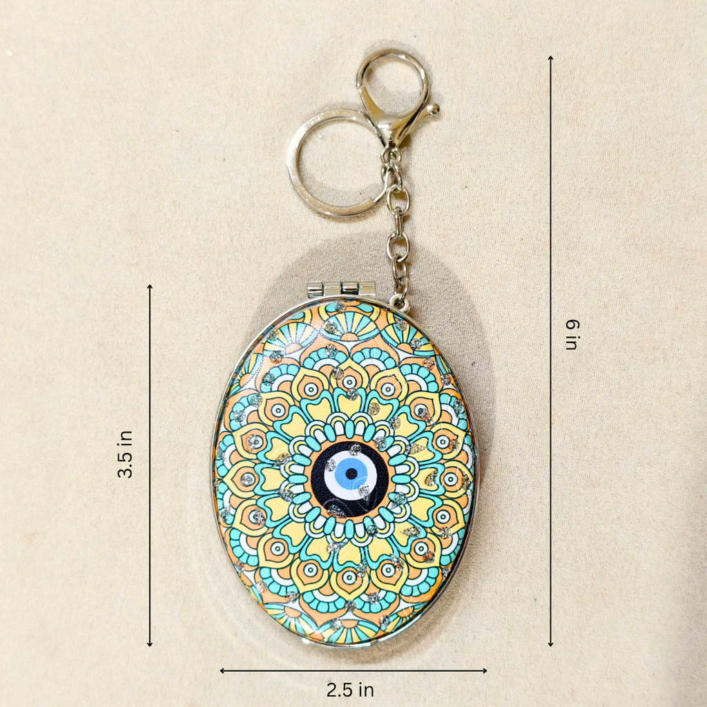 An oval keychain holder with a colorful mandala pattern and a blue-and-white evil eye in the center, shown on a beige background with dimension lines indicating 3.5 in height, 6 in overall height, and 2.