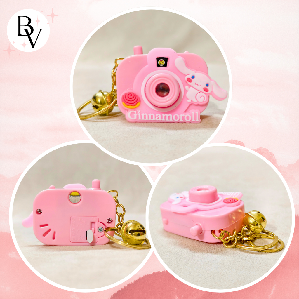 Three pink camera-shaped keychains with gold chains on a pink background, featuring the brand 'BV'.