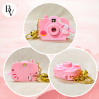 Three pink camera-shaped keychains with gold chains on a pink background, featuring the brand 'BV'.