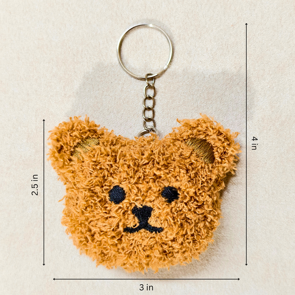 A plush keychain shaped like a cartoon Teddy face, featuring embroidered black eyes, and mouth details, a small nose, and a silver metal key ring and chain On a Beige Background.