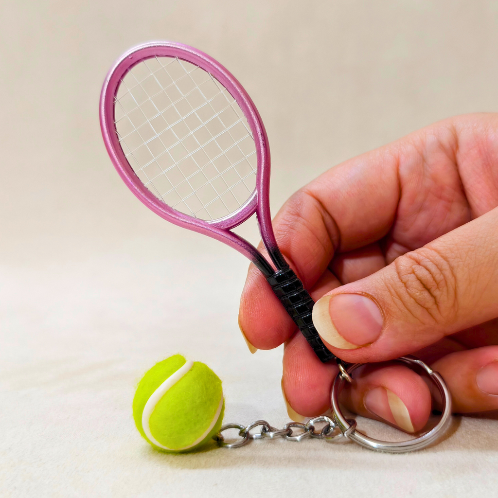A miniature pink tennis racket keychain with a small yellow tennis ball charm and silver metal ring, held by a hand against a light beige background.