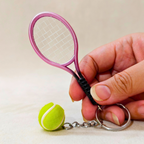 A miniature pink tennis racket keychain with a small yellow tennis ball charm and silver metal ring, held by a hand against a light beige background.