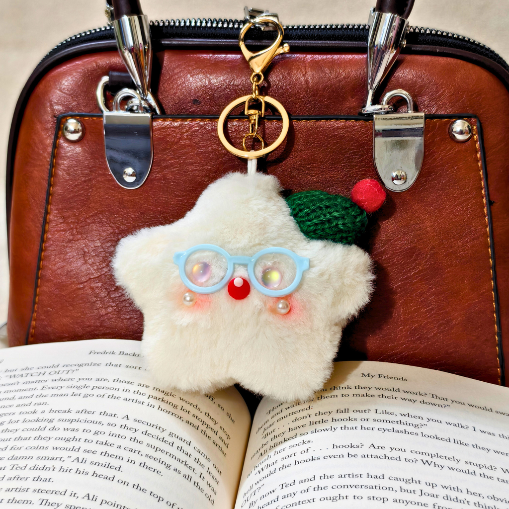 A fluffy white Santa Claus keychain charm with blue glasses, a green hat, and a red nose, hanging from a gold clasp on a brown leather handbag over an open book.