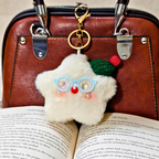 A fluffy white Santa Claus keychain charm with blue glasses, a green hat, and a red nose, hanging from a gold clasp on a brown leather handbag over an open book.