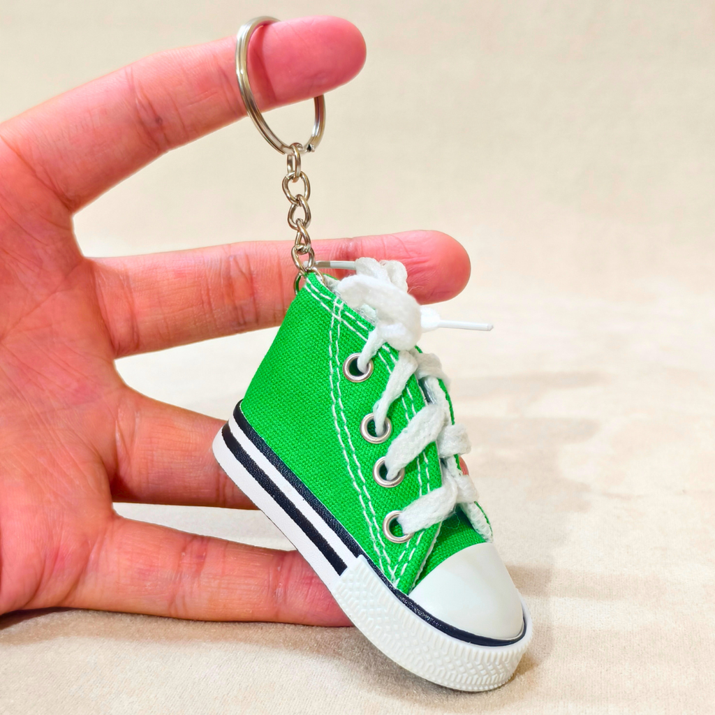 A miniature green canvas high-top sneaker keychain held in a hand. The keychain features white laces, a white rubber toe cap, a white sole with a black stripe, and a silver metal key ring and chain.