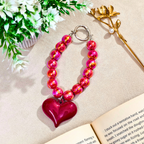 Heart-shaped pink and red beaded keychain on a beige surface with flowers and a book.
