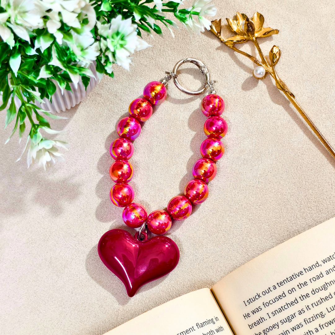Heart-shaped pink and red beaded keychain on a beige surface with flowers and a book.
