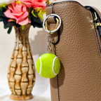 A tennis ball-shaped keychain charm with a silver split ring and chain, attached to a brown textured handbag. A decorative vase with pink flowers is blurred in the background.