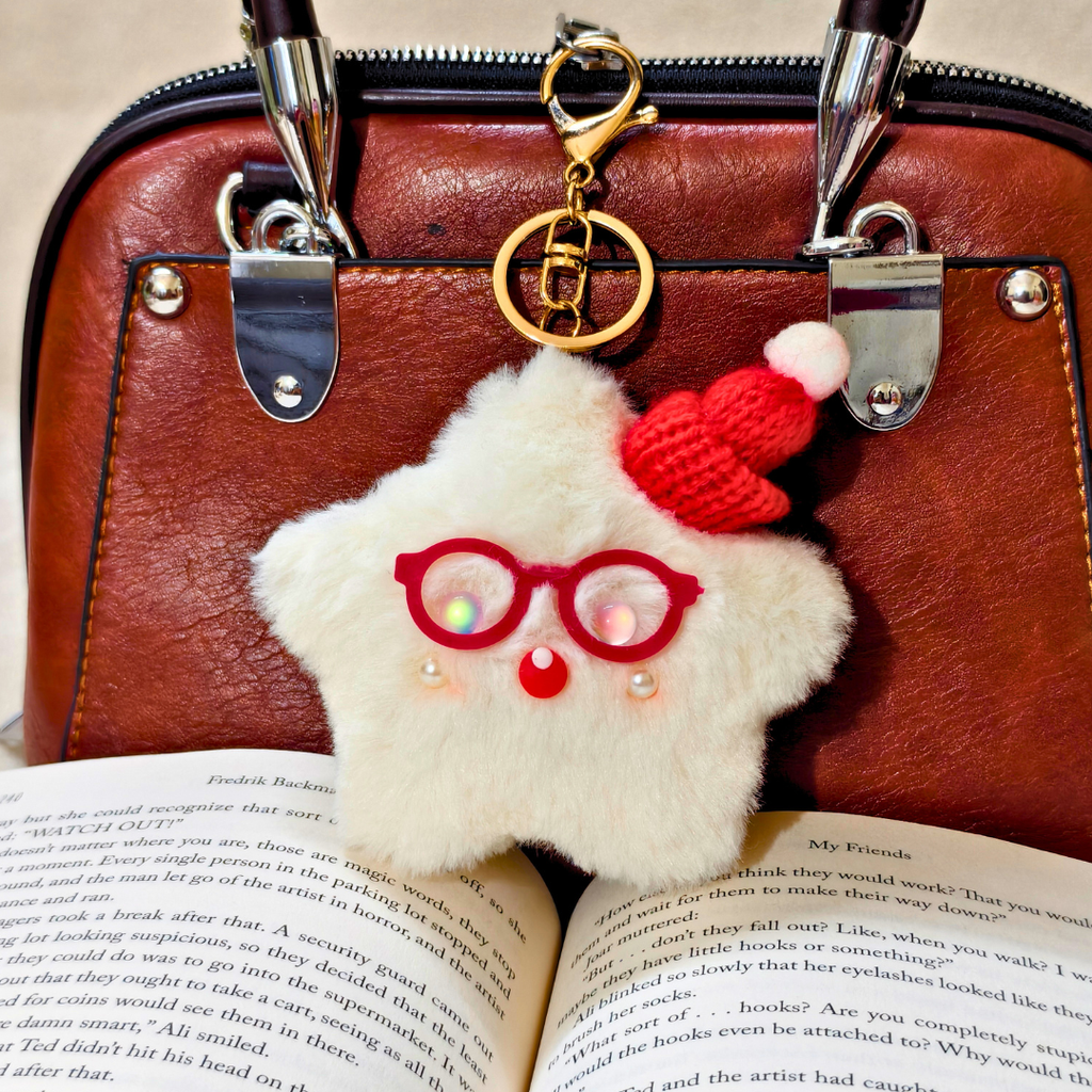 A fluffy white faux-fur handbag charm with red-rimmed glasses, a red knit hat, and pearl-like beads, hanging from a gold clasp on a brown leather handbag over an open book.