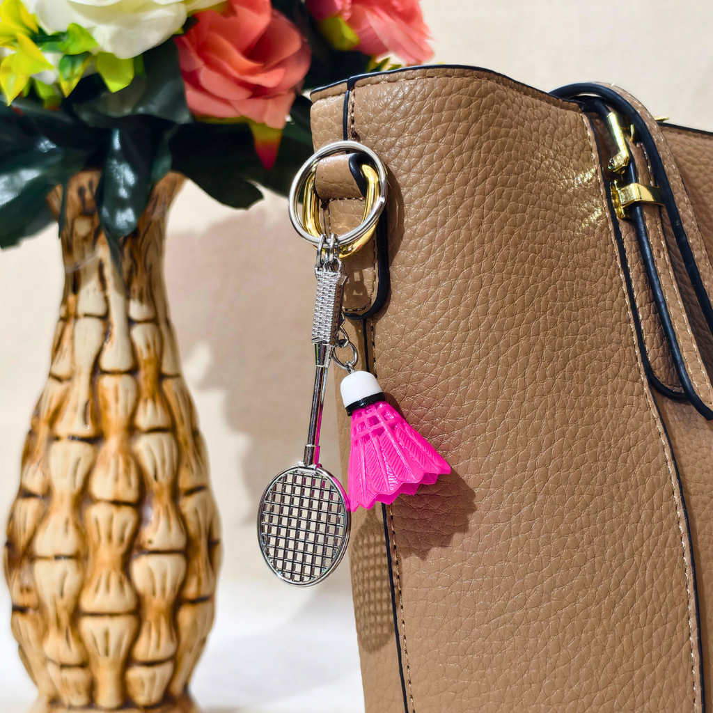  silver metal badminton racket keychain with a small pink and white plastic shuttlecock attached, Displayed on Tan handbag as bag charm against a plain beige background.