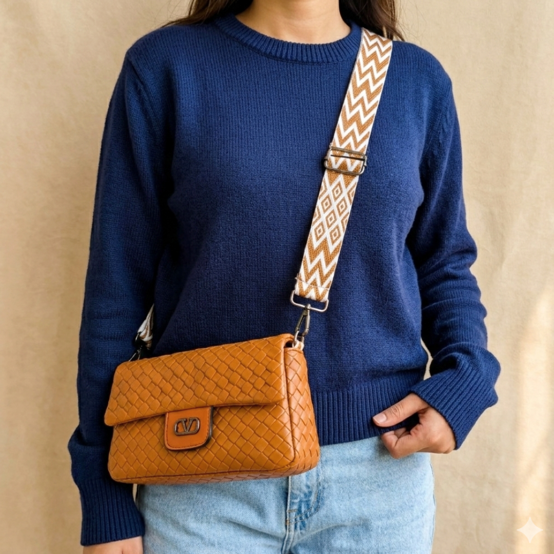 Person wearing a blue sweater with a brown leather bag and patterned strap against a beige background