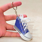 A miniature blue canvas high-top sneaker keychain with white laces and a silver metal key ring, held between a person's fingers against a beige background.