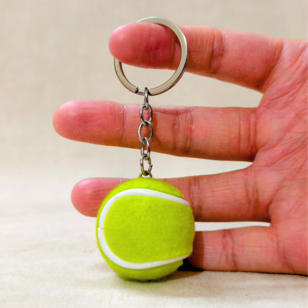 A miniature tennis ball keychain held between a person's fingers to show its small scale. The keychain features a bright yellow-green felt tennis ball charm with a white seam and a silver metal split ring and chain.