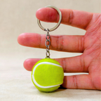 A miniature tennis ball keychain held between a person's fingers to show its small scale. The keychain features a bright yellow-green felt tennis ball charm with a white seam and a silver metal split ring and chain.