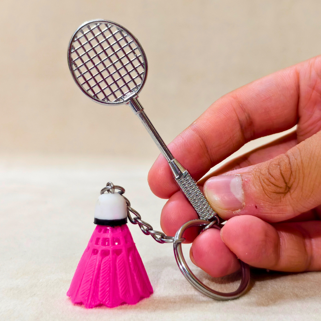 A hand holding a silver metal badminton racket keychain with a small pink and white plastic shuttlecock attached, against a plain beige background.