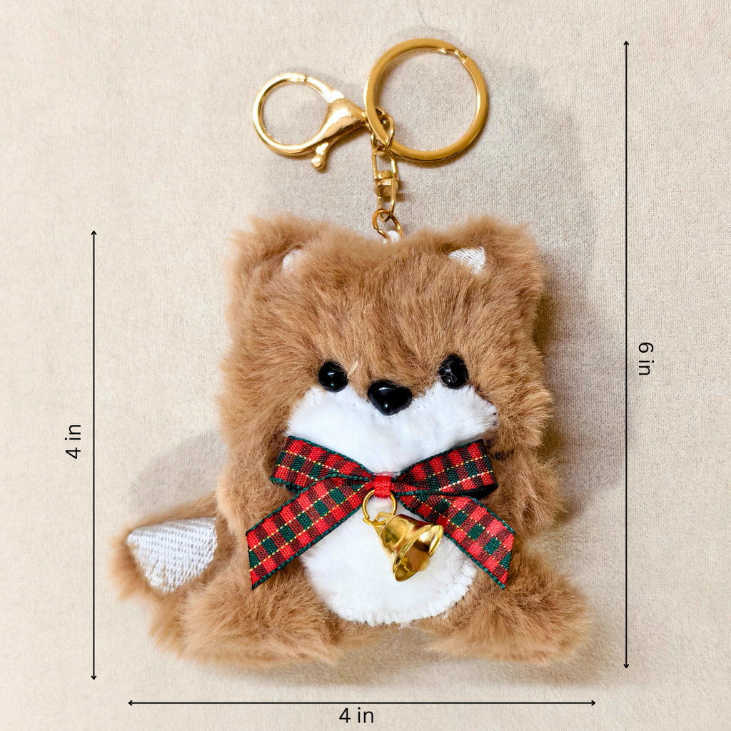 A plush brown dog keychain with a white snout and chest, black eyes and nose, a red and green plaid bow, and a small gold bell, shown on a beige background with dimension lines indicating 4 in by 6 in.