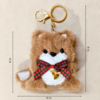 A plush brown dog keychain with a white snout and chest, black eyes and nose, a red and green plaid bow, and a small gold bell, shown on a beige background with dimension lines indicating 4 in by 6 in.
