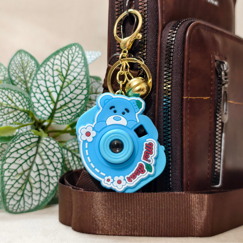 A blue cartoon bear-shaped camera keychain charm with gold hardware, shown attached to a brown leather bag next to a green leafy plant.