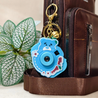 A blue cartoon bear-shaped camera keychain charm with gold hardware, shown attached to a brown leather bag next to a green leafy plant.
