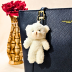 White teddy bear keychain attached to a black textured handbag with red roses in the background.