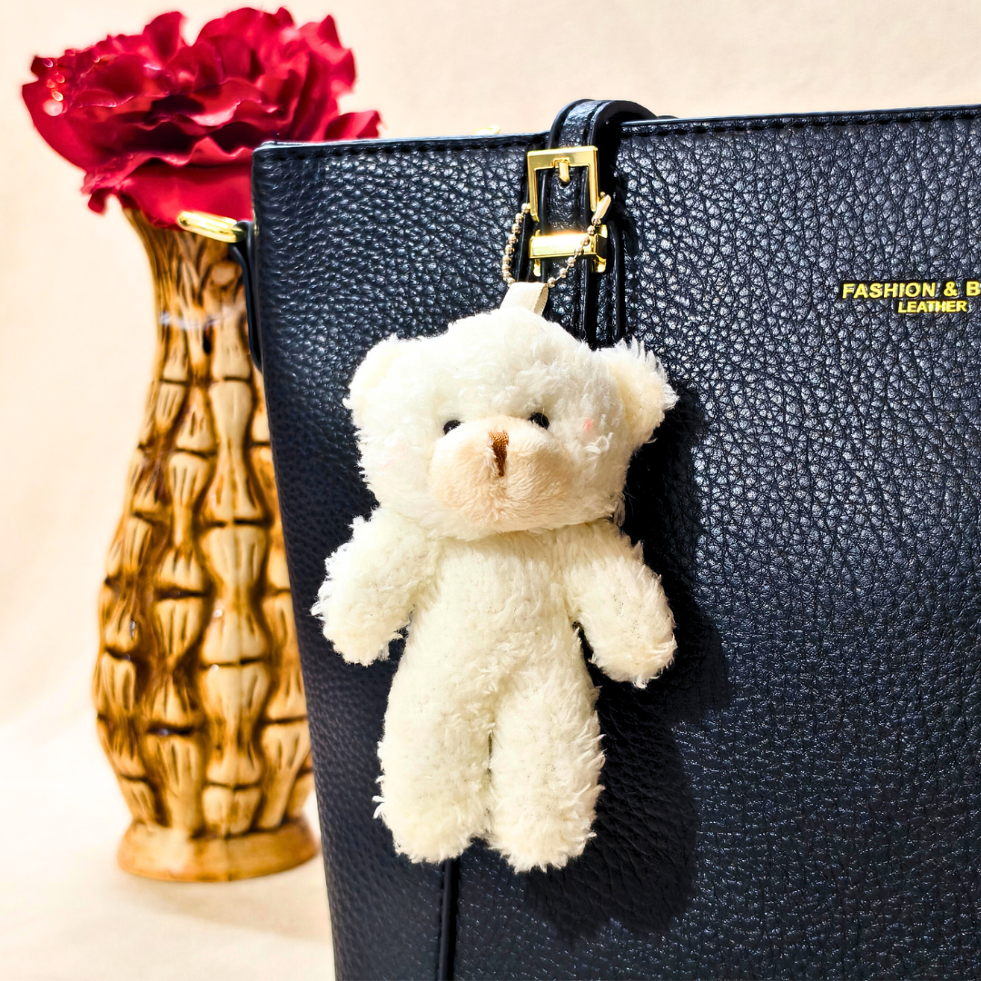 White teddy bear keychain attached to a black textured handbag with red roses in the background.