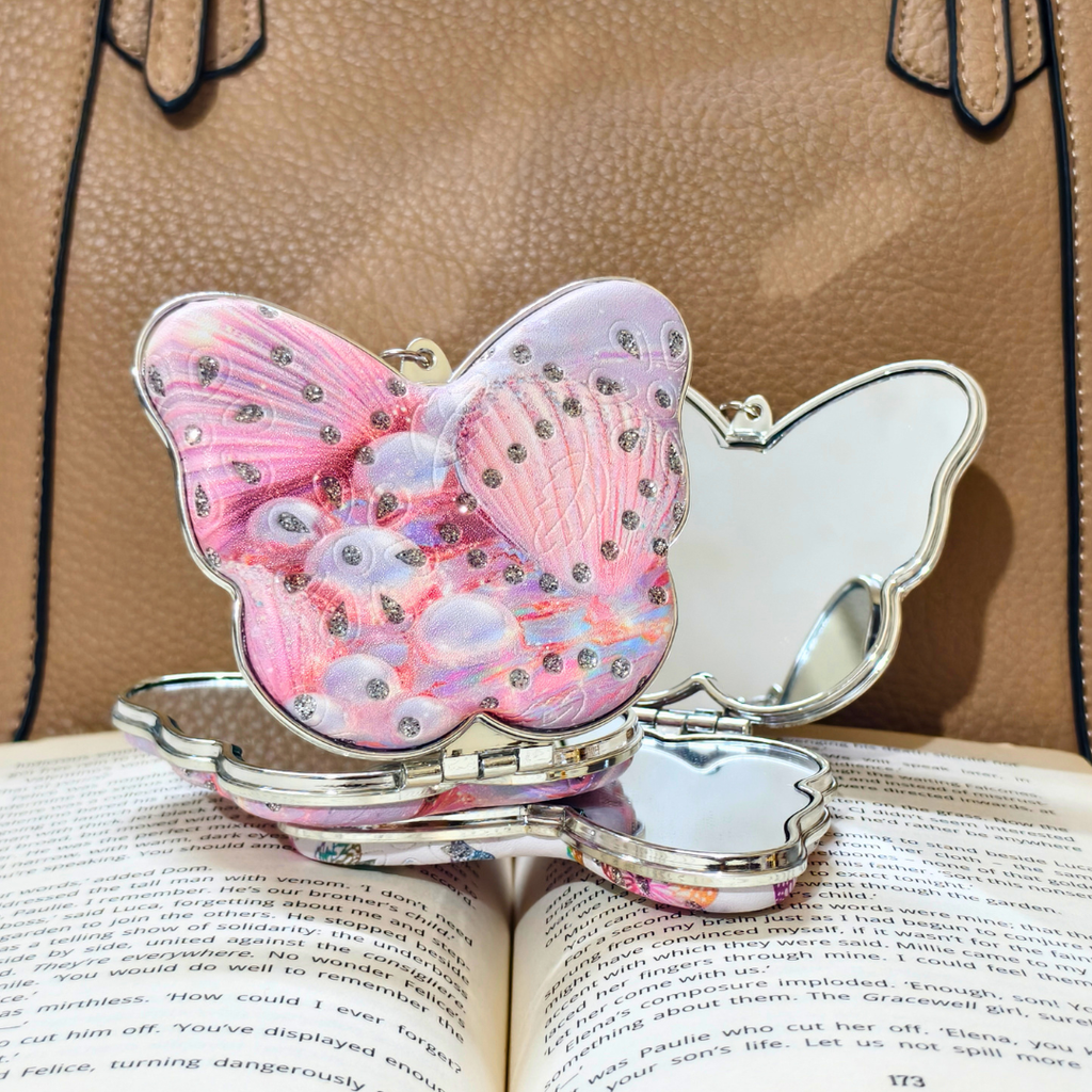 A butterfly-shaped compact mirror with a pink glittery wing design and rhinestone accents, resting open on an open book with a tan handbag in the background.