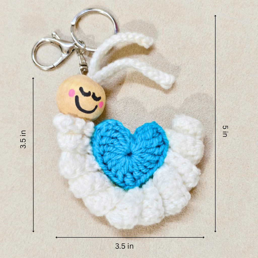 A crocheted angel keychain with white wings, a bright blue heart-shaped chest, and a wooden smiling face charm, shown on a beige background with black dimension lines and text indicating 3.5 in measurements.