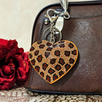Heart-shaped keychain with leopard print design attached to a brown leather bag with red roses in the background.