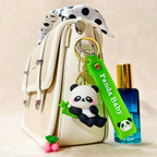 Panda keychain with a green strap labeled 'Panda Baby' attached to a beige bag, with a blue perfume bottle in the background.