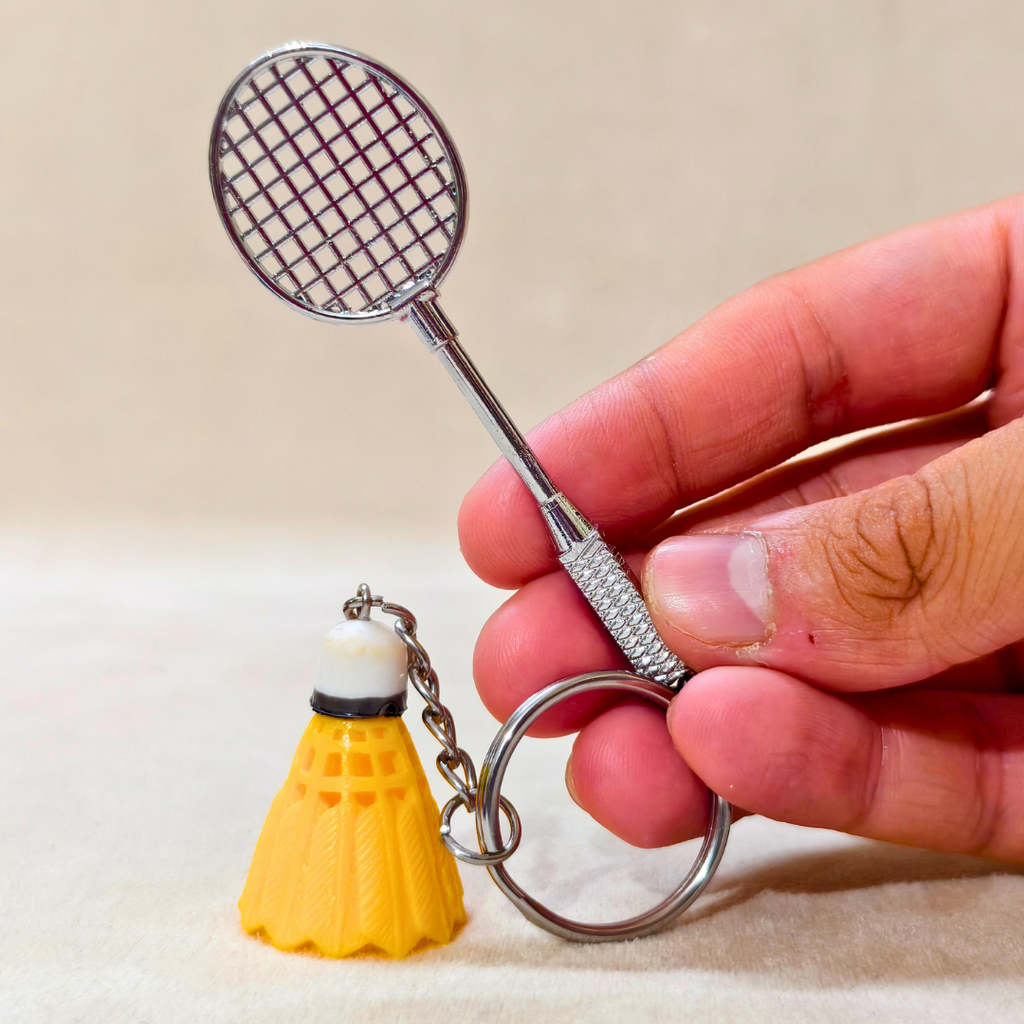 silver metal badminton racket keychain with a small pink and white plastic shuttlecock attached, holding in hand  against a plain beige background.


