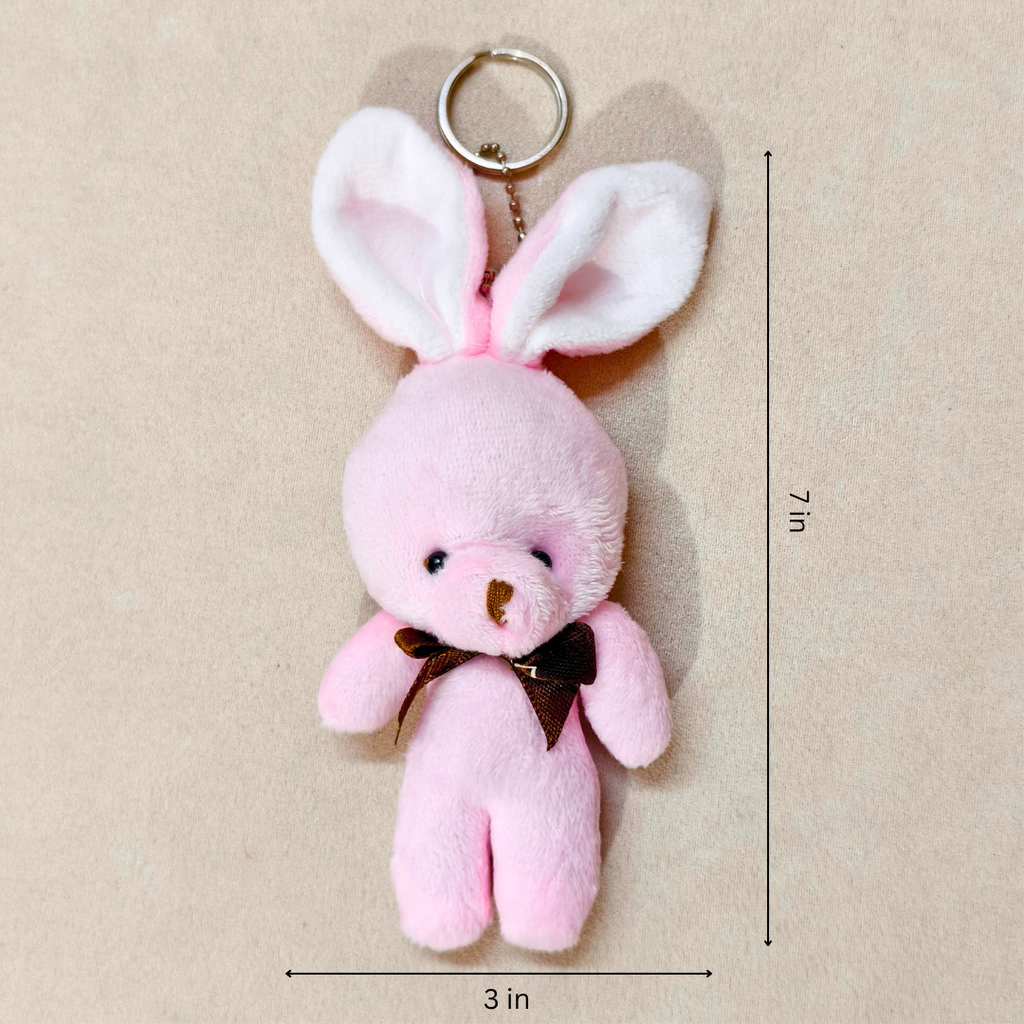 A pink plush bunny keychain with long floppy ears, a metal key ring, and a small dark brown bow at the neck, shown on a beige background with dimension lines indicating 3 inches wide by 7 inches tall.