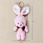 A pink plush bunny keychain with long floppy ears, a metal key ring, and a small dark brown bow at the neck, shown on a beige background with dimension lines indicating 3 inches wide by 7 inches tall.